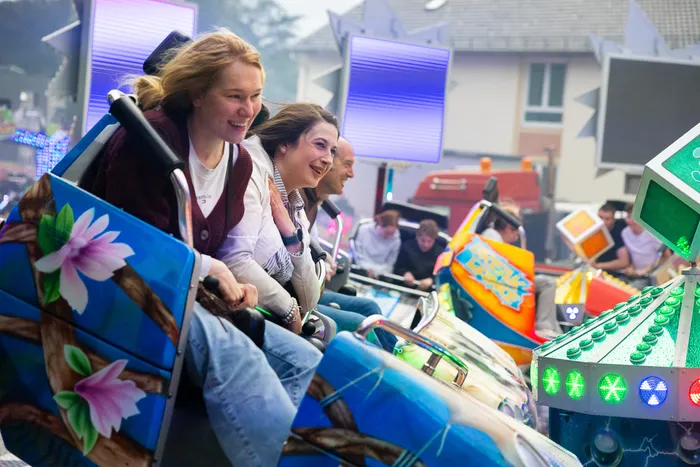 Zwei lächelnde Frauen sitzen in einer bunt dekorierten Fahrgeschäftskabine, während im Hintergrund weitere Menschen und Attraktionen der Kirmes zu sehen sind.