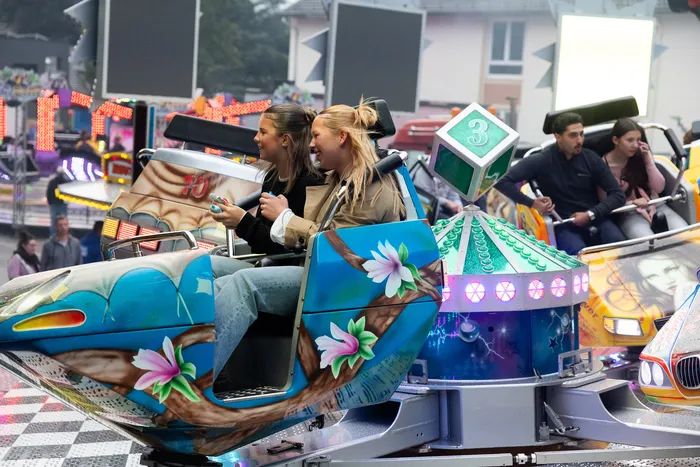Zwei junge Frauen sitzen lächelnd in einer blau und grün verzierten Karussell-Gondel. Im Hintergrund sind weitere Fahrgeschäfte und Besucher der Kirmes zu sehen.