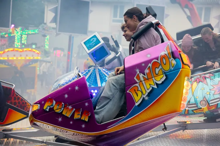 Junge Frau sitzt in einem farbenfrohen Karussellfahrzeug mit der Aufschrift „Bingo“ und spielt auf einem Bildschirm. Im Hintergrund sind weitere Fahrgeschäfte und Besucher der Kirmes zu sehen.