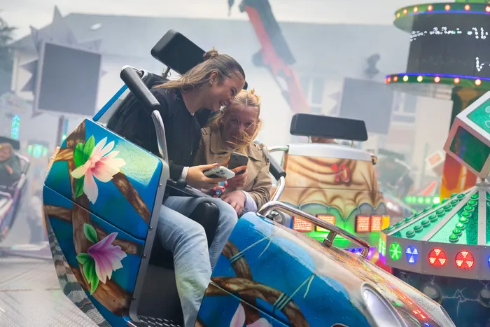 Zwei junge Erwachsene sitzen während der Kirmes in einer bunt dekorierten Fahrgeschäftskabine und schauen auf ein Smartphone.