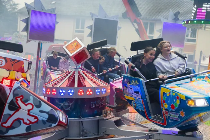 Kinder und Jugendliche fahren auf einem bunt geschmückten Karussell, im Hintergrund weitere Fahrgeschäfte und Buden einer Kirmes.