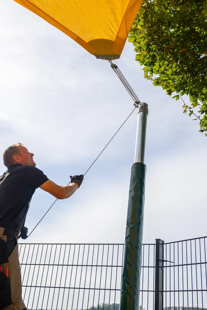 Ein Mann spannt ein gelbes Sonnensegel an einem grünen Mast fest.