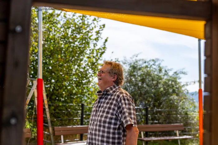 Ein lachender Mann mit Brille und kariertem Hemd steht unter einem gelben Sonnensegel.