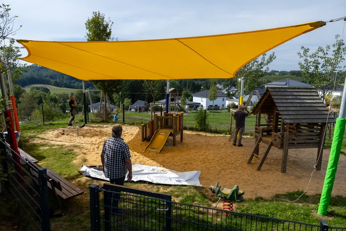 Ein großes gelbes Sonnensegel überspannt einen sandigen Spielplatz mit Spielgeräten und einem Holzhaus.