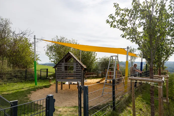 Ein Spielplatz mit Sandbereich, einem kleinen Holzhaus und einem großen gelben Sonnensegel, das über dem Spielbereich gespannt ist.