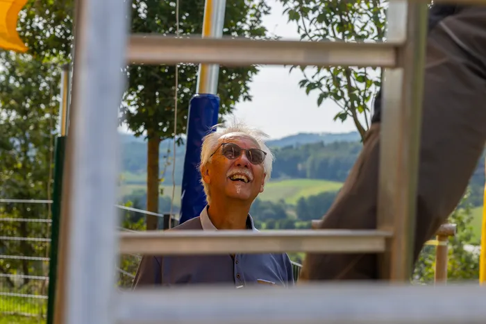 Ein lachender älterer Mann mit weißem Haar und Sonnenbrille blickt durch ein Metallgerüst nach oben.