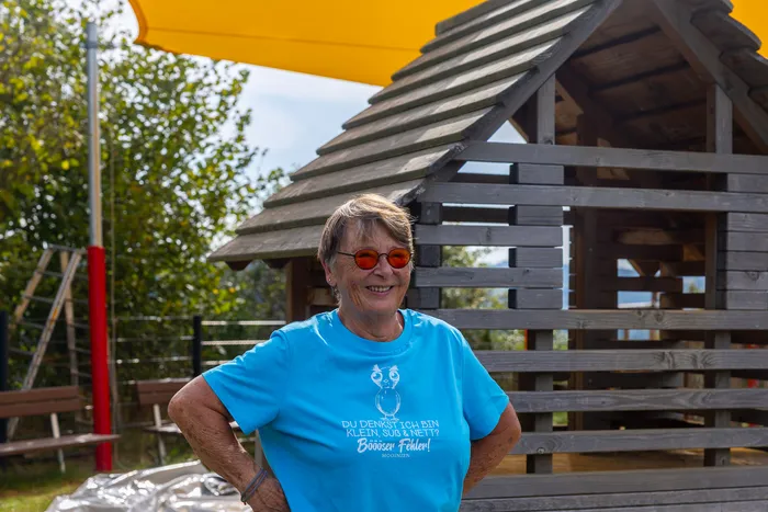 Eine Frau mit roter Sonnenbrille und blauem T-Shirt steht vor einem Holzspielhaus unter einem gelben Sonnensegel.