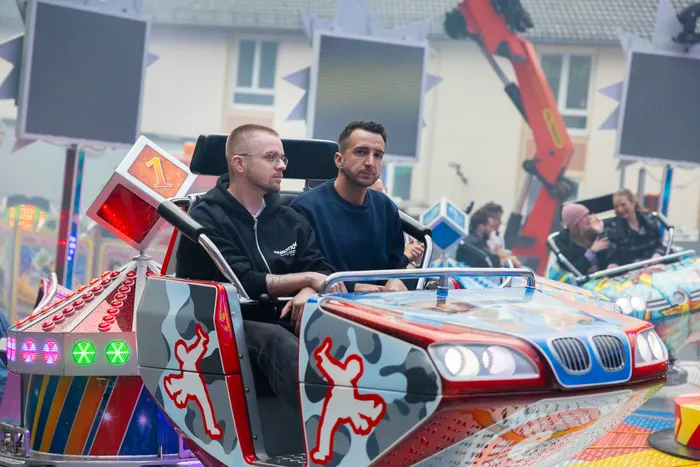Zwei junge Männer sitzen in Autoscootern auf einem Jahrmarkt. Im Hintergrund sind weitere Fahrgeschäfte und Besucher zu sehen.