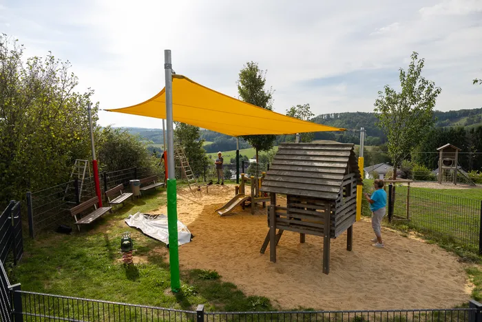 Ein Spielplatz mit Sandbereich, Holzspielhaus und Rutsche unter einem großen gelben Sonnensegel.