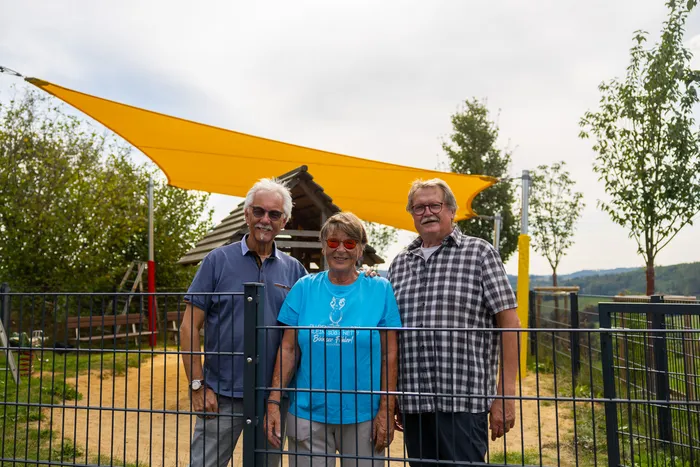 Drei Personen stehen vor einem großen gelben Sonnensegel über einem Spielplatz.