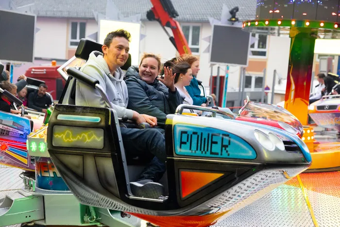 Drei junge Erwachsene sitzen lachend in einem Auto auf einer Fahrgeschäftsplattform, umgeben von weiteren Fahrgeschäften und Besuchern auf der Kirmes.