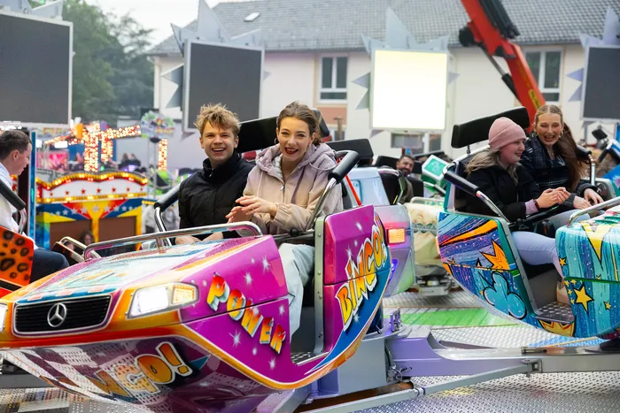 Junge Erwachsene lachen und fahren auf einem bunt dekorierten Karussell auf der Kirmes. Im Hintergrund sind weitere Fahrgeschäfte und Menschen zu sehen.