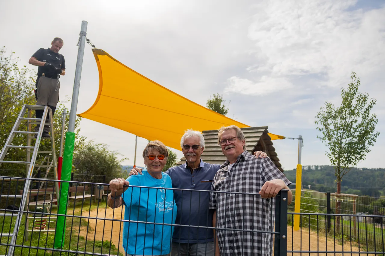 Drei Personen stehen vor einem großen gelben Sonnensegel auf einem Spielplatz, während im Hintergrund ein Mann auf einer Leiter an einem Mast arbeitet.