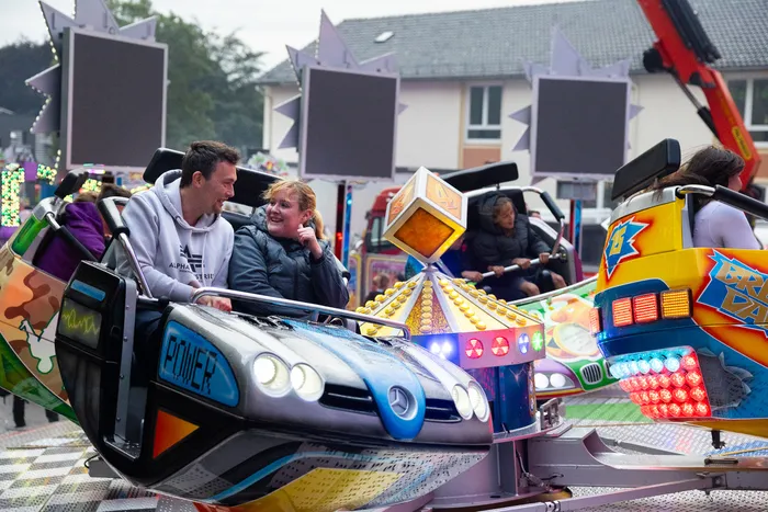 Ein Paar sitzt lachend in einem bunt dekorierten Karussellfahrzeug auf der Kirmes, im Hintergrund weitere Fahrgeschäfte und Besucher.