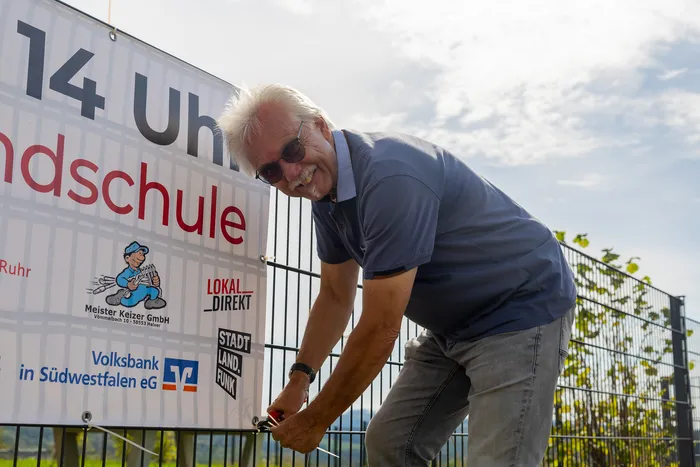 Ein lächelnder Mann mit weißen Haaren und Sonnenbrille arbeitet an einem großen Schild neben einem Metallzaun.