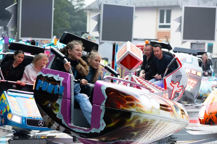 Zwei Mädchen sitzen lachend in einem bunt dekorierten Karussellwagen, umgeben von weiteren Fahrgästen auf der Kirmes.