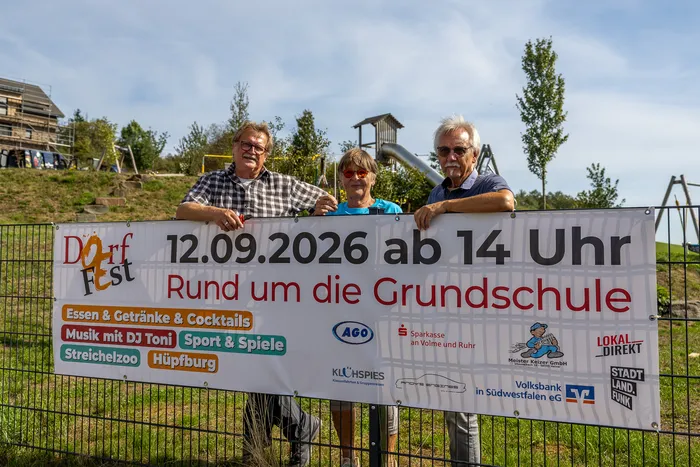 Drei Männer halten ein Banner für das Dorffest am 12. September 2026 rund um die Grundschule in Oberbrügge.