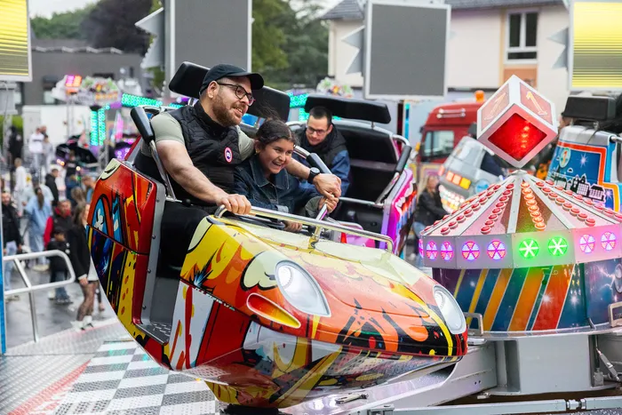 Mann und Teenager sitzen lachend in einer farbenfrohen Achterbahn auf der Kirmes, umgeben von Menschen und weiteren Fahrgeschäften.