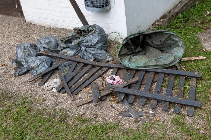 Schwarze Müllsäcke, ein grüner Sack und zerbrochene Holzlatten liegen auf dem Boden neben einer weißen Wand.