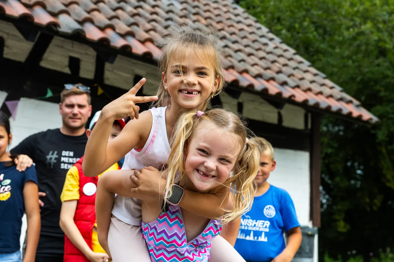 Zwei lachende Mädchen posieren gemeinsam, eines zeigt ein Victory-Zeichen. Im Hintergrund stehen weitere Personen vor einem Gebäude mit Ziegeldach.