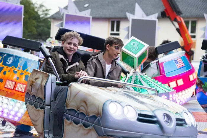 Zwei junge Männer sitzen in einem bunt dekorierten Karussellfahrzeug, das einem Auto nachempfunden ist. Im Hintergrund sind weitere Fahrgeschäfte und Menschen auf der Kirmes zu sehen.