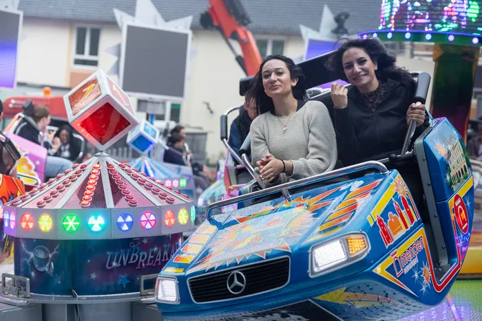 Zwei junge Frauen lächeln in einem blau-gelben Karussellfahrzeug, das wie ein Auto gestaltet ist. Im Hintergrund sind weitere Fahrgeschäfte und Menschen auf der Kirmes zu sehen.