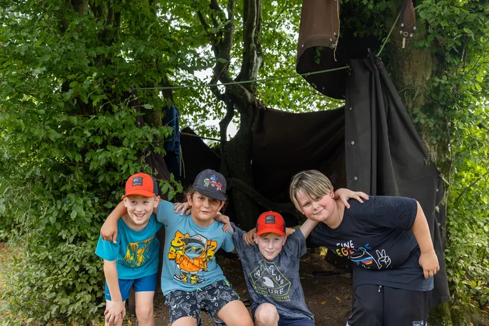 Vier lächelnde Kinder posieren vor einem improvisierten Zelt aus dunklem Stoff unter einem Baum.