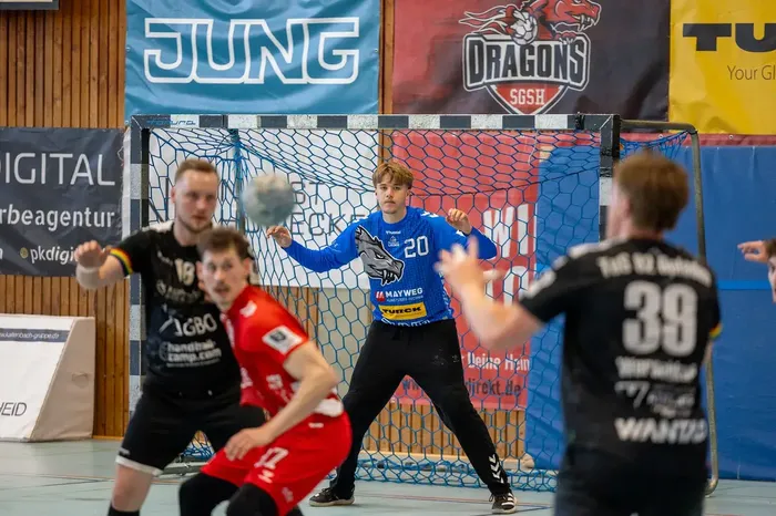 Handballspieler in blauer Ausrüstung springt zum Wurf vor dem Tor. Im Hintergrund sind weitere Spieler und Werbebanner zu sehen.