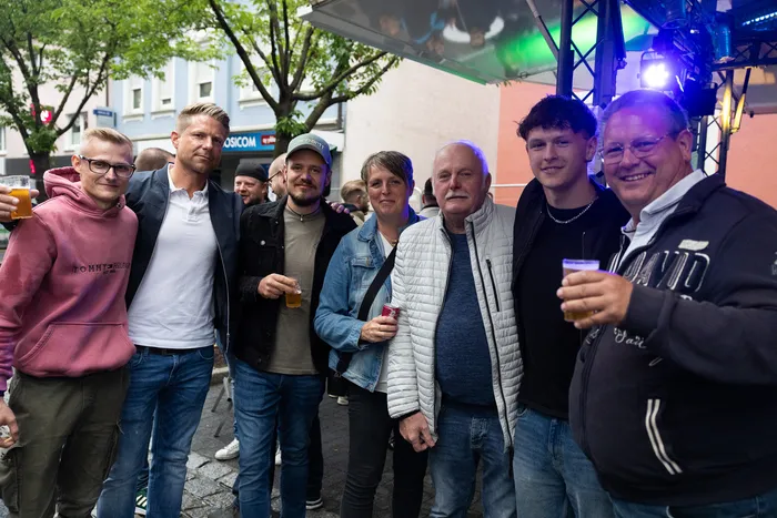 Eine Gruppe von Menschen unterschiedlichen Alters steht eng zusammen und hält Biergläser hoch, im Hintergrund eine festlich beleuchtete Bühne.