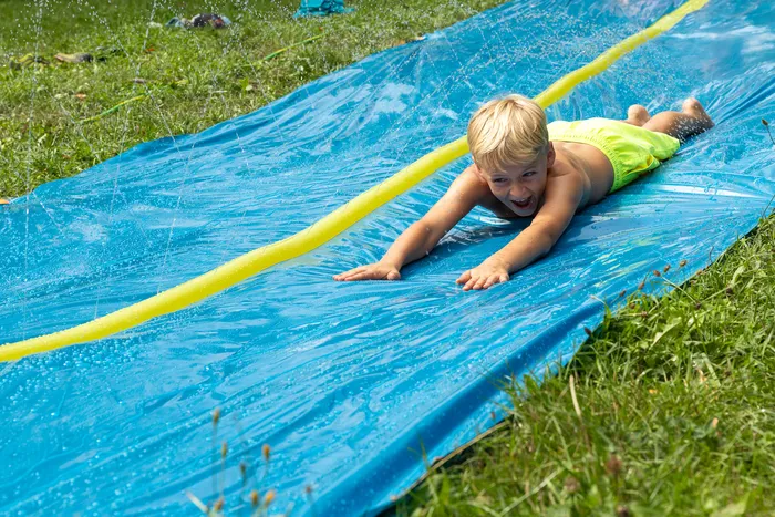 Ein lachender Junge in gelber Badehose rutscht auf einer mit Wasser bespritzten blauen Plane über eine Wiese.