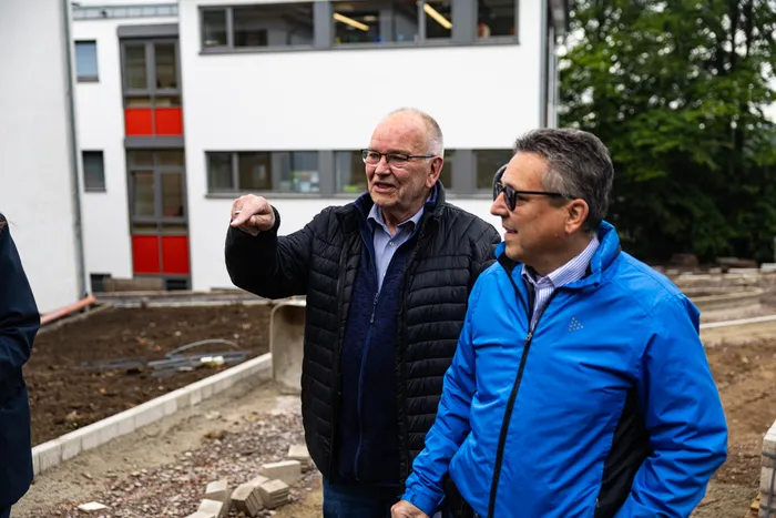 Zwei Männer mittleren Alters stehen vor einem modernen Schulgebäude auf einer Baustelle. Der Mann links zeigt mit ausgestreckter Hand nach links, während der Mann rechts ihn aufmerksam betrachtet.