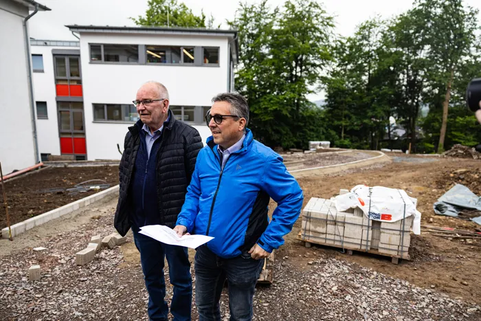 Zwei Männer stehen auf einer Baustelle vor einem Gebäude mit roten und grauen Fassadenelementen. Im Vordergrund liegt ein Bauplan, rechts sind gestapelte Baumaterialien zu sehen.