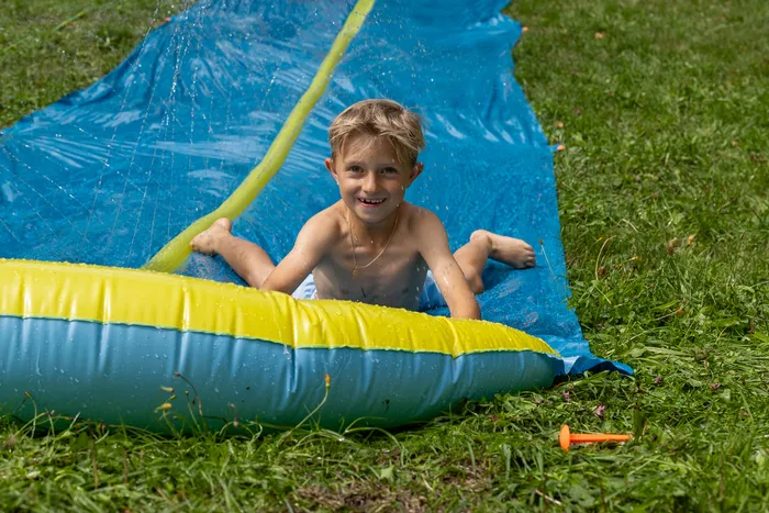 Ein lächelnder Junge spielt auf einer blauen aufblasbaren Wasserrutsche auf einer Wiese.