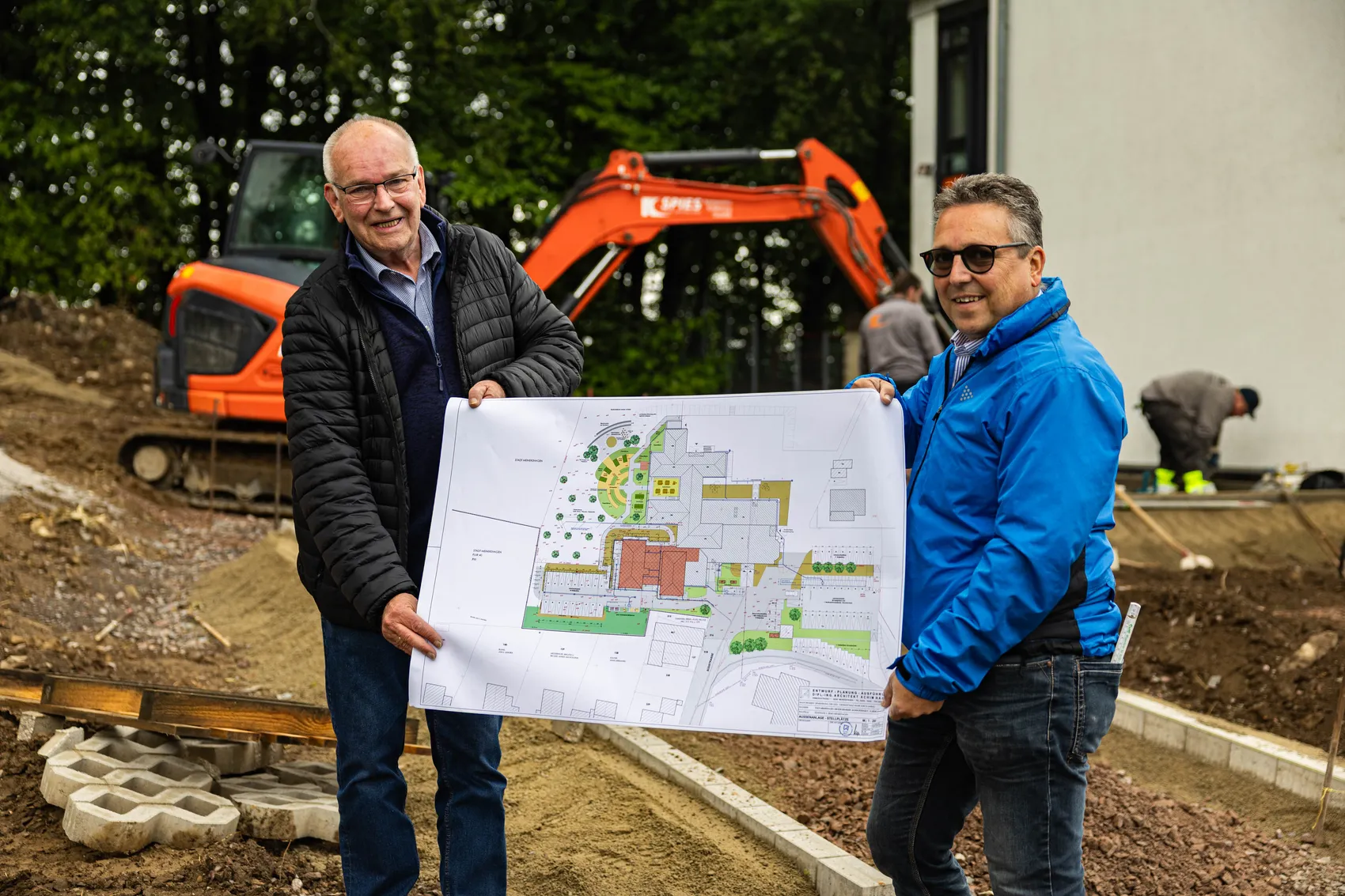Zwei Männer halten einen Bauplan vor einer Baustelle mit Baggern. Im Hintergrund ist ein Gebäude und Grün zu sehen.