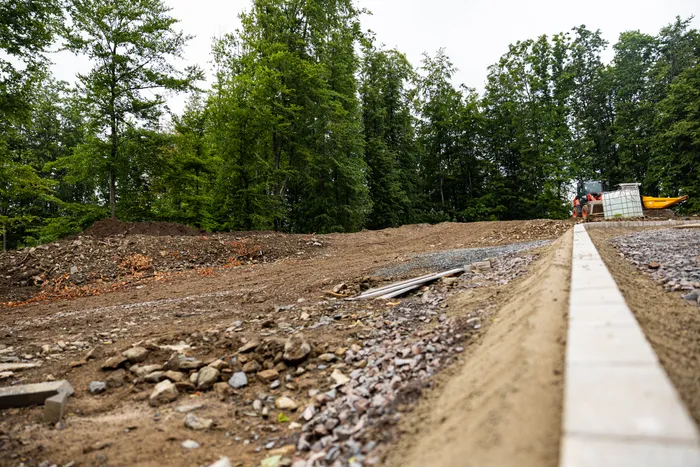 Eine Baustelle mit ausgehobenem Erdreich und Kiesflächen vor einem Wald. Im Hintergrund ist ein Bagger zu sehen. Der neue Schulhof der Grundschule auf der Wahr in Meinerzhagen befindet sich im Umbau.