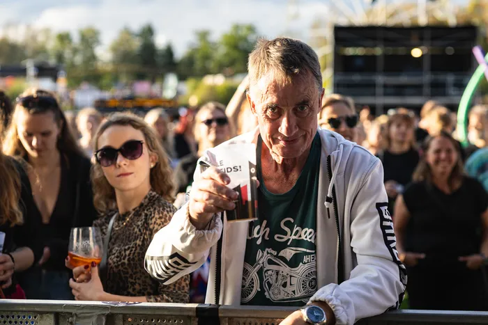 Ein Mann in weißer Jacke und grünem T-Shirt hält ein kleines Glas in die Kamera, umgeben von einer Menschenmenge beim Bautz-Festival.