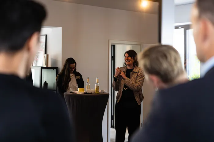 Eine Frau mit gelocktem Haar spricht enthusiastisch mit gestikulierenden Händen vor einer Gruppe von Menschen, die um einen Tisch mit Getränken stehen. Im Hintergrund ist ein Raum mit moderner Einrichtung zu sehen.