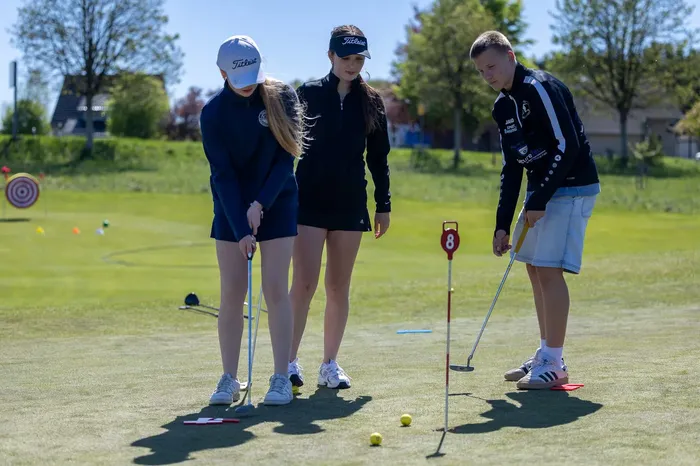 Drei Jugendliche stehen auf einem Golfplatz und bereiten sich auf einen Putt vor. Sie tragen Sportkleidung und schauen auf den Golfball. Im Hintergrund ist eine grüne Wiese und ein Ziel zu sehen.