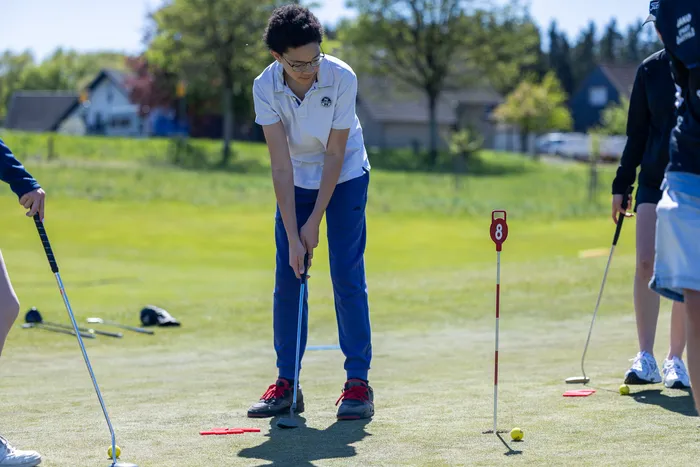 Ein Jugendlicher mit dunklen lockigen Haaren puttet auf einem Golfplatz, während andere Spieler im Hintergrund stehen.