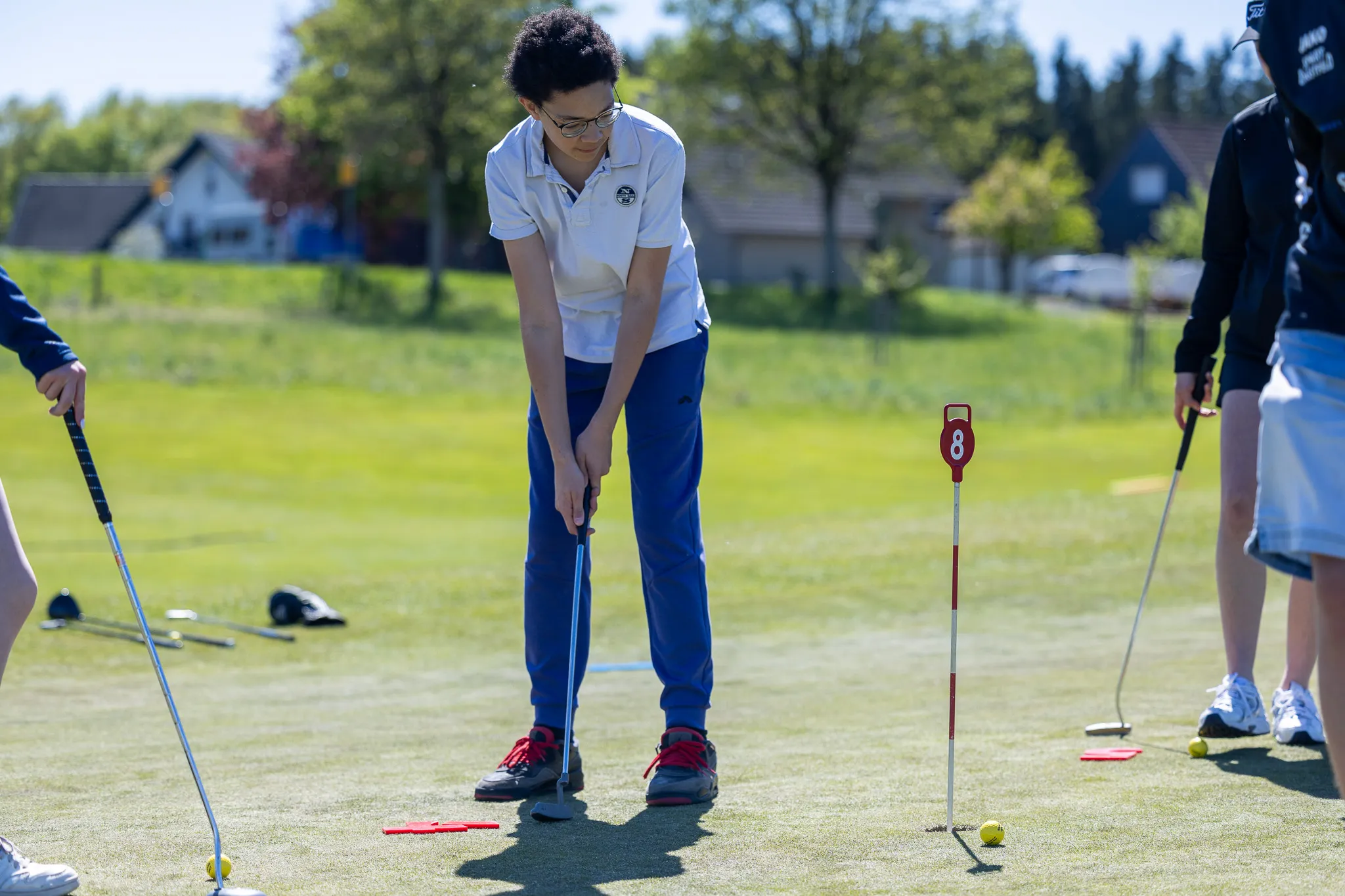 Ein Jugendlicher mit dunklen lockigen Haaren puttet auf einem Golfplatz, während andere Spieler im Hintergrund stehen.