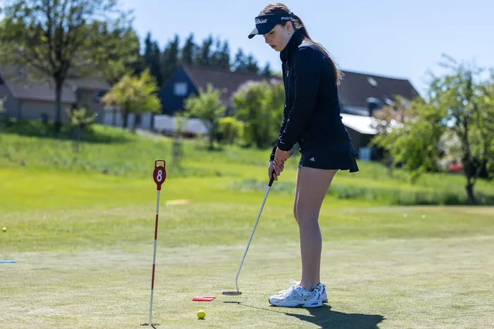 Eine junge Golferin konzentriert sich auf das Putten auf einem Golfplatz. Im Hintergrund sind Häuser und Bäume zu sehen.