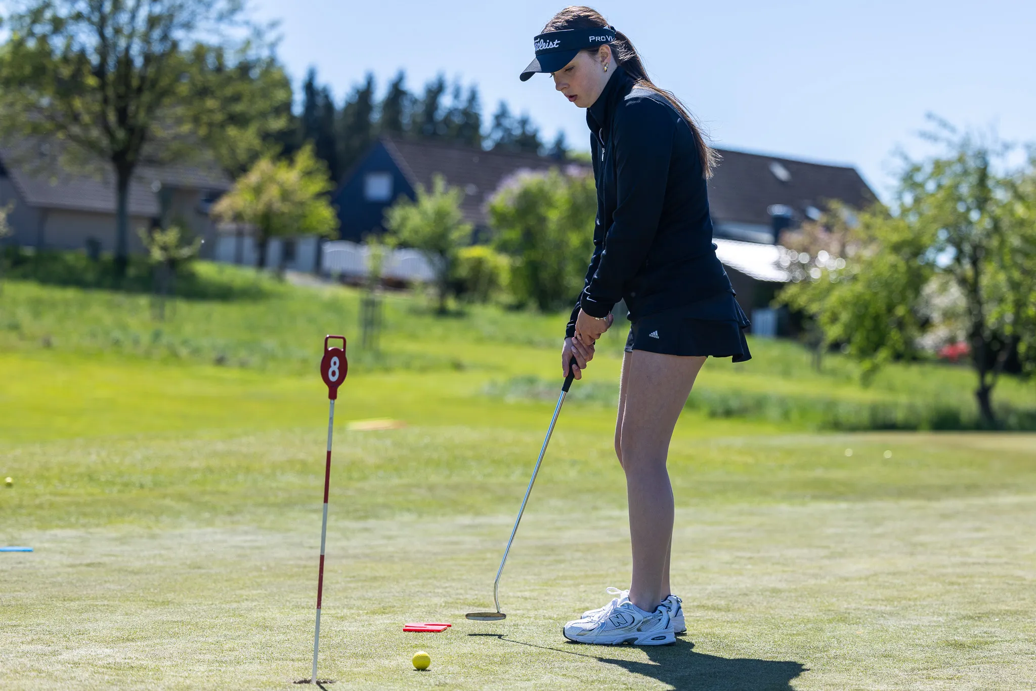 Eine junge Golferin konzentriert sich auf das Putten auf einem Golfplatz. Im Hintergrund sind Häuser und Bäume zu sehen.