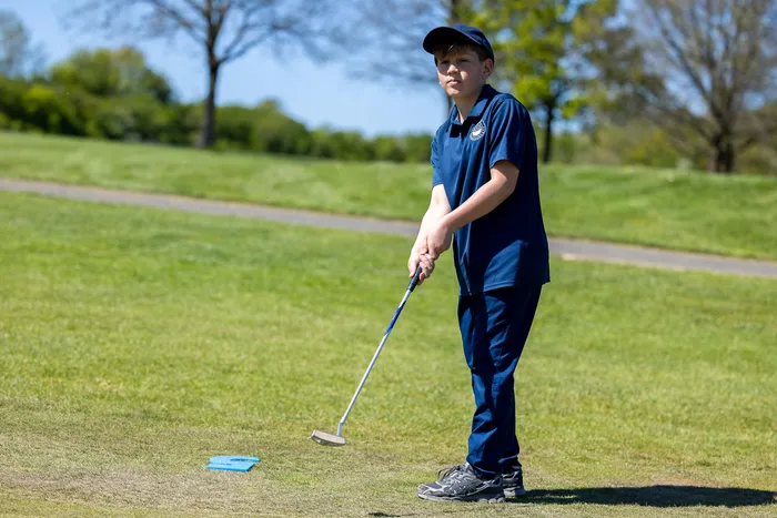 Ein Jugendlicher mit Golfschläger steht auf einem Golfplatz und bereitet einen Schlag vor. Im Hintergrund eine grüne Landschaft und ein Weg.