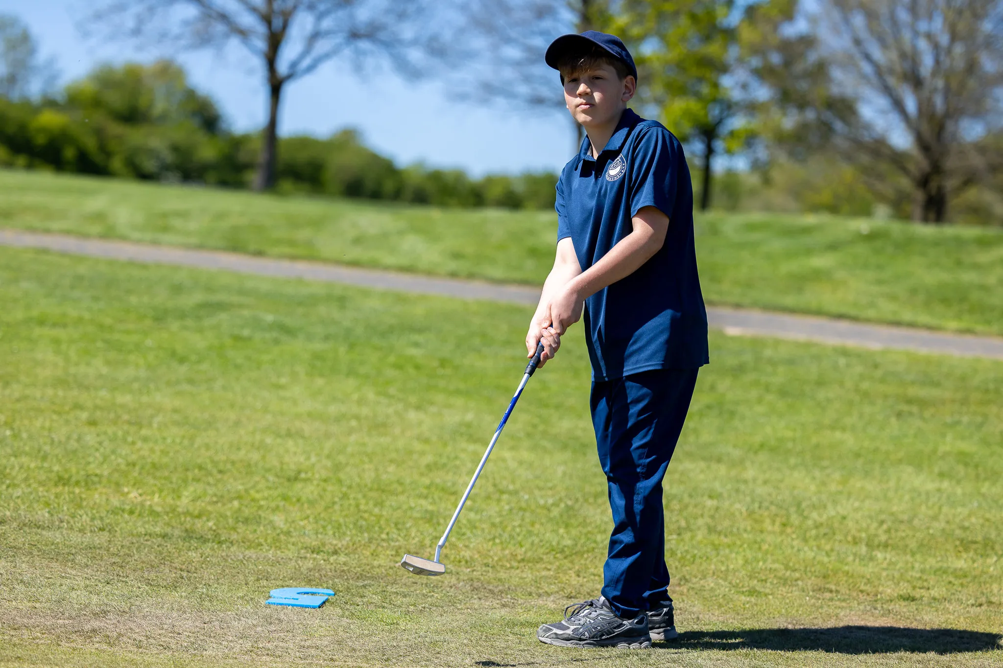 Ein Jugendlicher mit Golfschläger steht auf einem Golfplatz und bereitet einen Schlag vor. Im Hintergrund eine grüne Landschaft und ein Weg.