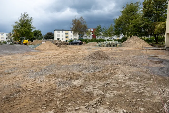 Baustelle mit Sand- und Kieshaufen, einem gelben Radlader und einem schwarzen Auto vor Wohngebäuden.