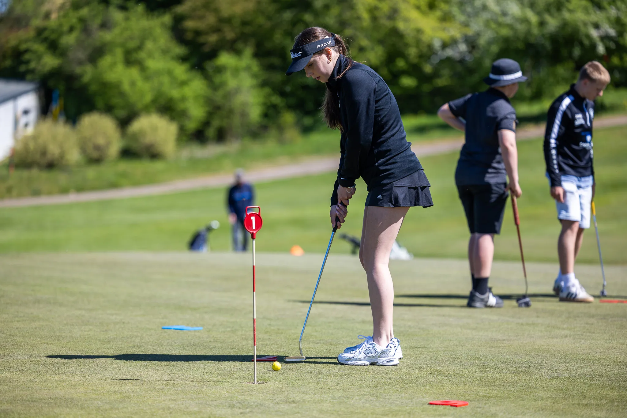 Schülerin puttet auf dem Golfplatz. Im Hintergrund weitere Golfer und das Grün des Golfclubs Dreibäumen.