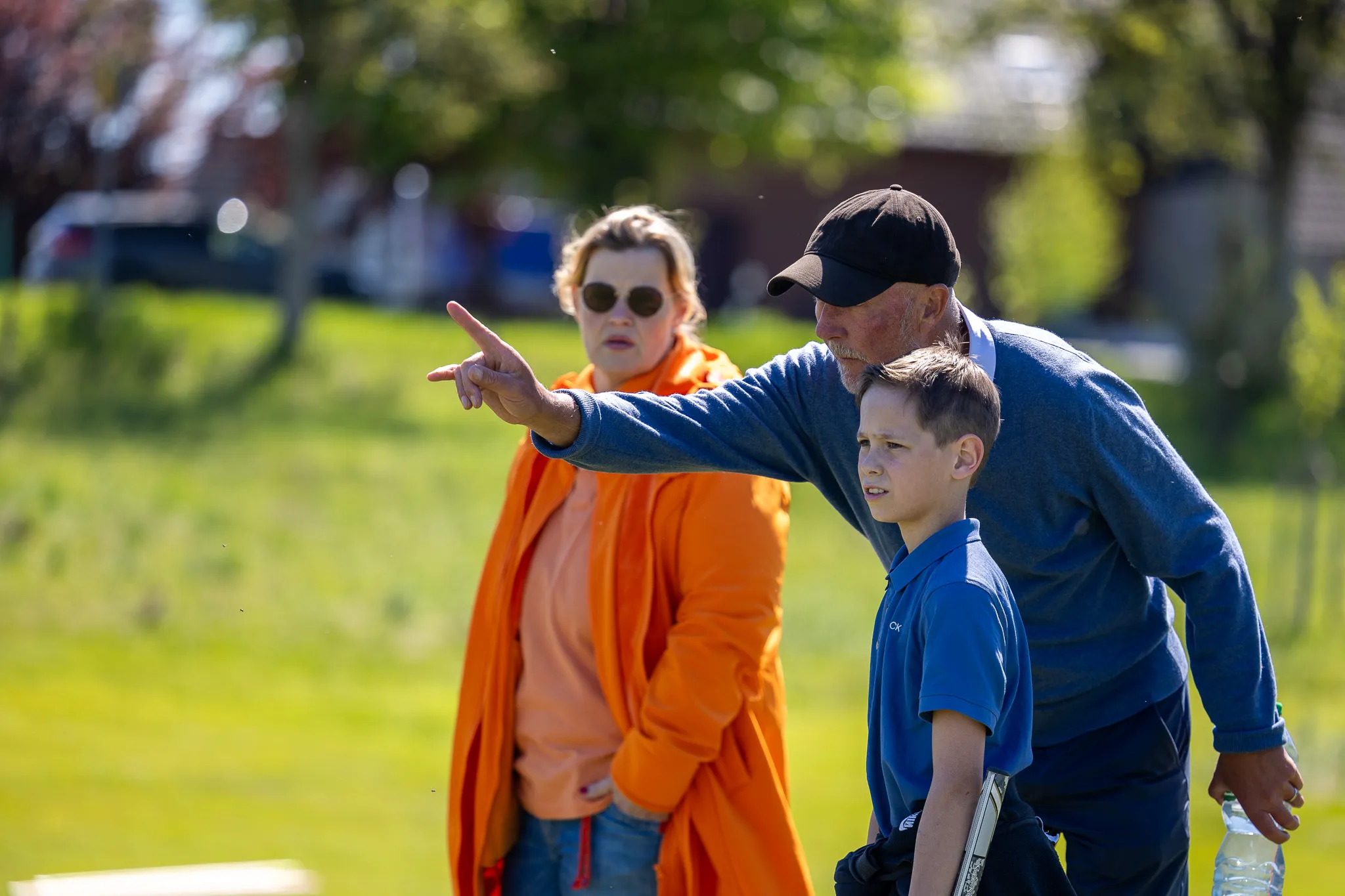 Ein Mann zeigt einem Jungen mit Golfschläger die Richtung, während eine Frau im Hintergrund zuschaut. Im Hintergrund sind Bäume und ein Golfplatz zu sehen.