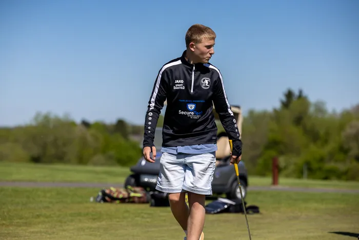 Ein junger Mann mit blonden Haaren geht auf einem Golfplatz, einen Schläger in der Hand tragend. Er trägt ein blaues Trainingsshirt mit Werbeaufdruck und weiße Shorts.