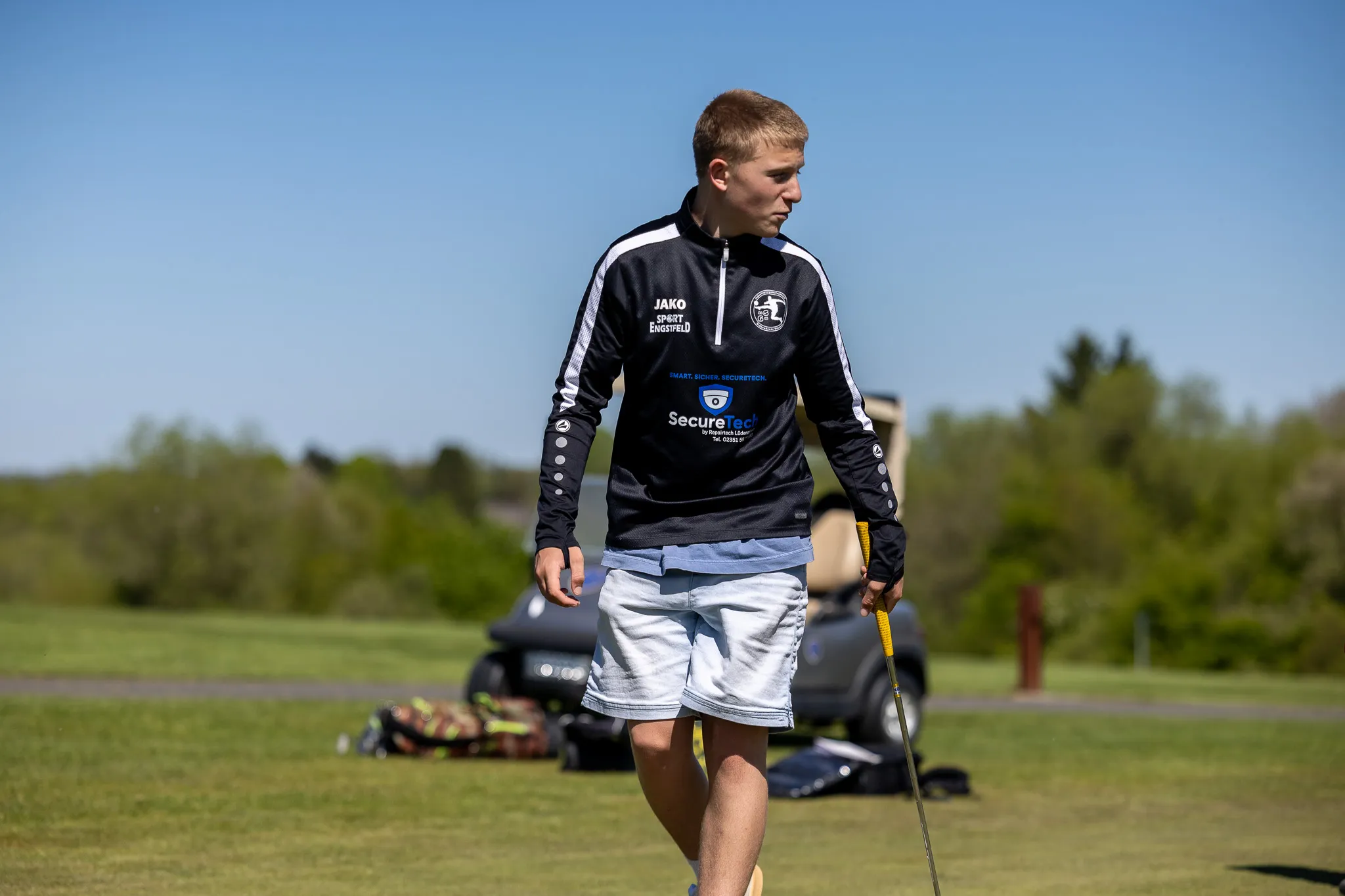 Ein junger Mann mit blonden Haaren geht auf einem Golfplatz, einen Schläger in der Hand tragend. Er trägt ein blaues Trainingsshirt mit Werbeaufdruck und weiße Shorts.