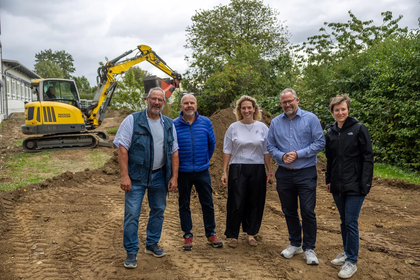 Fünf Personen stehen auf der Baustelle des Schulhofs der Regenbogenschule vor einem gelben Bagger.